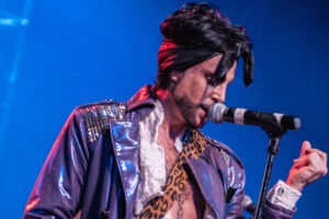 A man in purple jacket and leopard print hat singing.